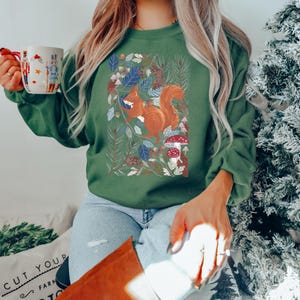 May include: A green sweatshirt featuring a graphic of a squirrel in a woodland scene. The squirrel is brown with a fluffy tail. The woodland is depicted with green leaves, pine cones, and mushrooms.