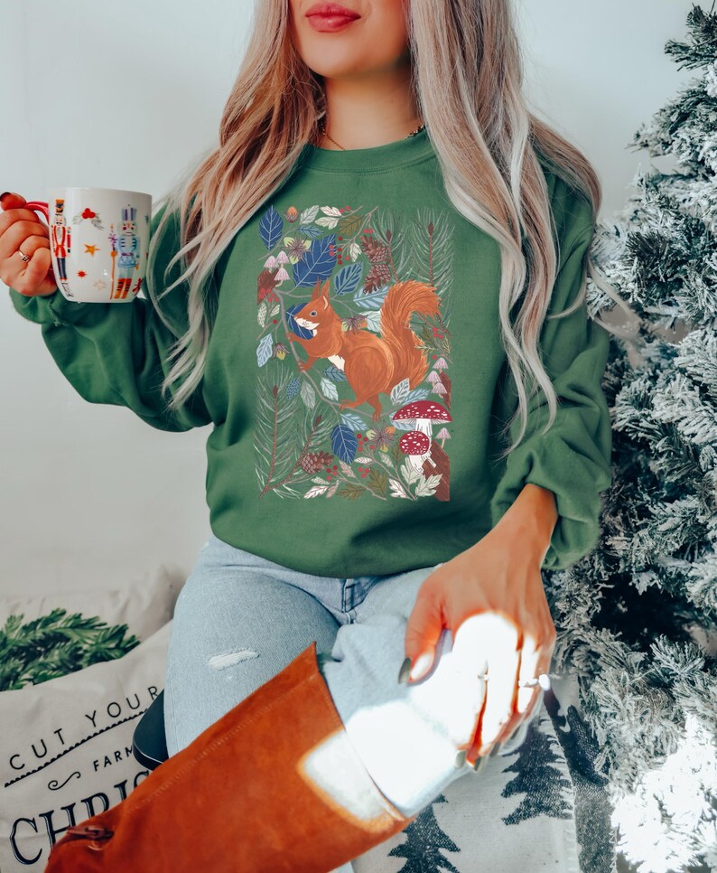May include: A green sweatshirt featuring a graphic of a squirrel in a woodland scene. The squirrel is brown with a fluffy tail. The woodland is depicted with green leaves, pine cones, and mushrooms.
