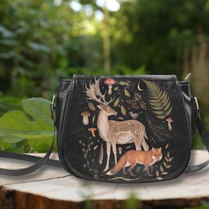 Op de afbeelding: Zwarte crossbodytas met een illustratie met een natuurthema. Het ontwerp toont een hert, een vos, paddenstoelen en bladeren. De tas heeft een gebogen vorm, een lange riem en een ritssluiting.