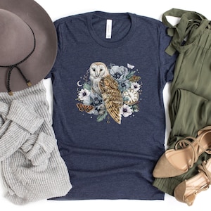 May include: A navy blue t-shirt with a white barn owl illustration surrounded by flowers, butterflies, and a vintage pocket watch. The owl has a white face and brown and white feathers.
