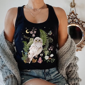 May include: Black vest top with a white owl illustration surrounded by ferns, flowers, and crystals. A crescent moon and stars are also depicted. The top is sleeveless and made of a soft material.