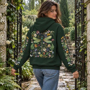 May include: A dark forest green hooded sweatshirt featuring a large, detailed graphic on the back of a dragonfly surrounded by various colorful wildflowers, ferns, and small butterflies. The sleeves also feature matching floral patterns.