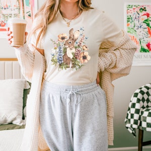 Op de afbeelding: Een cr&egrave;mekleurig T-shirt met een afbeelding van een bruin konijn omringd door kleurrijke bloemen. Het konijn kijkt naar links. Het shirt wordt gedragen door een persoon met lang bruin haar.