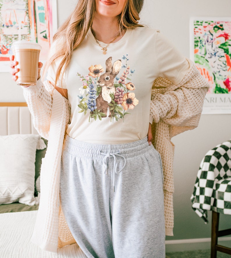 Op de afbeelding: Een cr&egrave;mekleurig T-shirt met een afbeelding van een bruin konijn omringd door kleurrijke bloemen. Het konijn kijkt naar links. Het shirt wordt gedragen door een persoon met lang bruin haar.