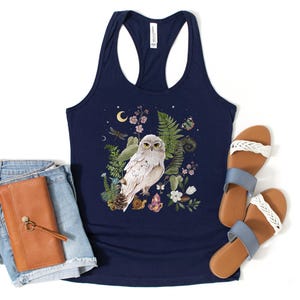 May include: Navy blue vest top with a detailed illustration of a white owl surrounded by ferns, flowers, and crystals. The design includes a crescent moon and a dragonfly. The image also shows denim shorts, a brown wallet, and sandals.