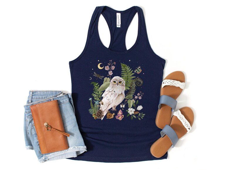 May include: Navy blue vest top with a detailed illustration of a white owl surrounded by ferns, flowers, and crystals. The design includes a crescent moon and a dragonfly. The image also shows denim shorts, a brown wallet, and sandals.
