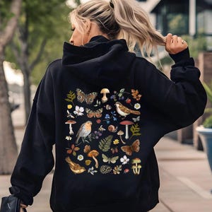 Pode incluir: Moletom preto com um design temático da natureza nas costas. O design inclui borboletas, pássaros, cogumelos, flores e folhagens. O moletom tem capuz e mangas compridas.