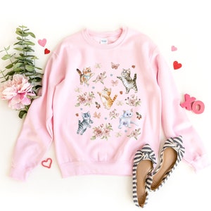 May include: A pink sweatshirt with a graphic print of multiple cats and flowers. The cats are in various poses, including sitting, standing, and jumping. The flowers are in shades of pink, purple, and white.