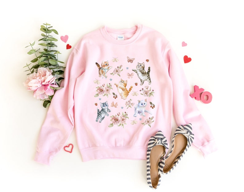 May include: A pink sweatshirt with a graphic print of multiple cats and flowers. The cats are in various poses, including sitting, standing, and jumping. The flowers are in shades of pink, purple, and white.