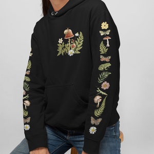 May include: Black hoodie with a nature-themed design. The front features a mushroom, snail, and floral arrangement. Sleeves are adorned with illustrations of mushrooms, butterflies, flowers, and a frog. The hoodie has a drawstring hood and a front pocket.