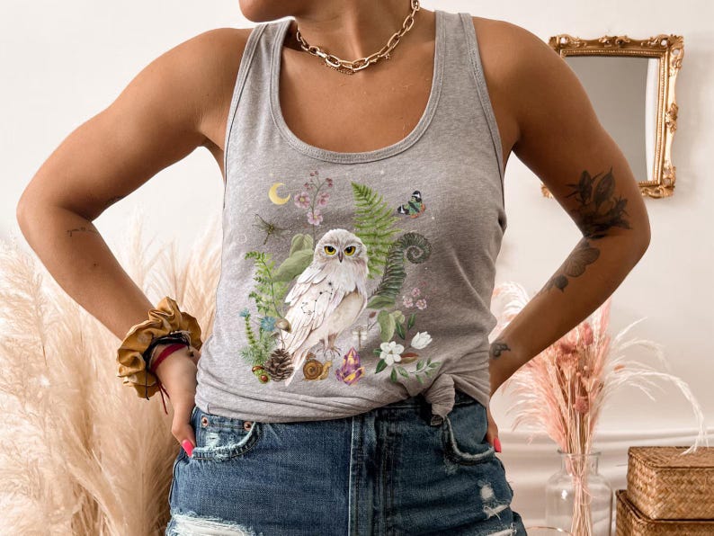 May include: Grey vest top with a detailed illustration of a white owl surrounded by ferns, flowers, and crystals. A crescent moon and dragonfly are also depicted. The design has a whimsical, nature-inspired theme.