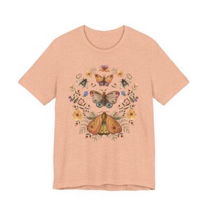 May include: A peach-colored short-sleeve t-shirt featuring a central graphic print of three moths arranged vertically, surrounded by a circular wreath of various small flowers, leaves, and sprigs in muted tones of yellow, blue, and green.