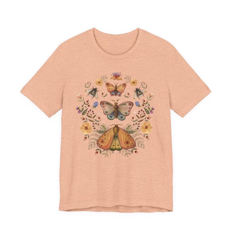 May include: A peach-colored short-sleeve t-shirt featuring a central graphic print of three moths arranged vertically, surrounded by a circular wreath of various small flowers, leaves, and sprigs in muted tones of yellow, blue, and green.
