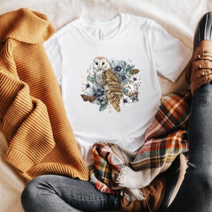May include: A white t-shirt with a graphic of a barn owl with blue and purple flowers and a clock. The owl is white with brown markings. The flowers are watercolor style.