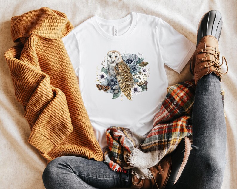 May include: A white t-shirt with a graphic of a barn owl with blue and purple flowers and a clock. The owl is white with brown markings. The flowers are watercolor style.