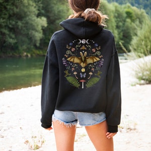 May include: Black hooded sweatshirt with a graphic print of a bat surrounded by flowers, mushrooms, and other woodland elements. The graphic is centred on the back of the sweatshirt.