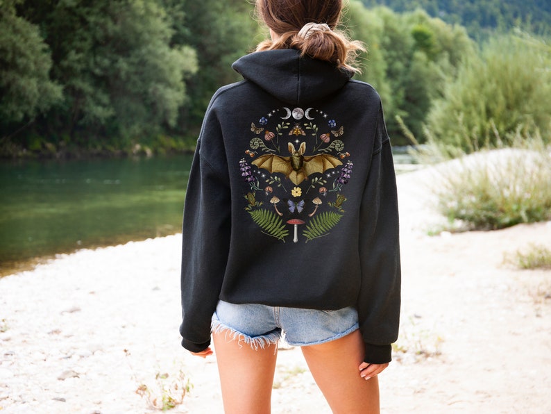 May include: Black hooded sweatshirt with a graphic print of a bat surrounded by flowers, mushrooms, and other woodland elements. The graphic is centred on the back of the sweatshirt.