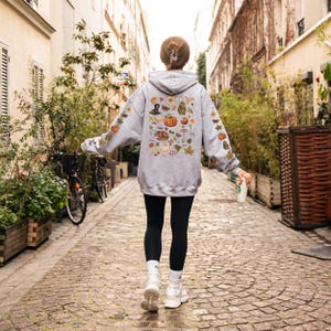 Puede incluir: Una sudadera gris con un estampado colorido de tem&aacute;tica oto&ntilde;al en la espalda. El estampado presenta calabazas, hojas, bellotas y otros elementos oto&ntilde;ales.