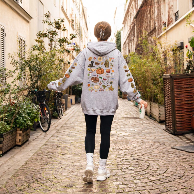 Puede incluir: Una sudadera gris con un estampado colorido de tem&aacute;tica oto&ntilde;al en la espalda. El estampado presenta calabazas, hojas, bellotas y otros elementos oto&ntilde;ales.
