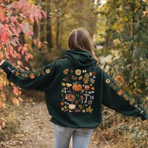 Puede incluir: Una sudadera con capucha verde oscuro con un gran estampado de elementos oto&ntilde;ales en la espalda. El estampado incluye calabazas, hojas, setas, bellotas, un zorro, un b&uacute;ho y un girasol. La sudadera tiene capucha y bolsillo canguro.
