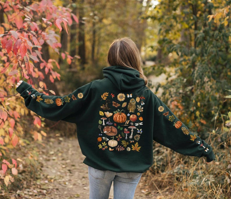 Puede incluir: Una sudadera con capucha verde oscuro con un gran estampado de elementos oto&ntilde;ales en la espalda. El estampado incluye calabazas, hojas, setas, bellotas, un zorro, un b&uacute;ho y un girasol. La sudadera tiene capucha y bolsillo canguro.