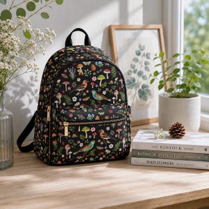 May include: Black backpack with a colorful pattern of birds, mushrooms, and flowers. The backpack has a top handle, adjustable straps, and a front zippered pocket. Gold-colored zippers and hardware.