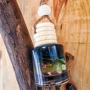 Puede incluir: Una pequeña botella de vidrio transparente con un líquido azul oscuro y flores secas en su interior. La botella está suspendida por una cuerda natural y detalles de madera. El producto está colgado de una rama de madera.