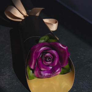 May include: A handcrafted, dark purple rose with green leaves emerges from a gold-colored, heart-shaped holder. The rose is part of a black, cylindrical package tied with a beige ribbon. The background is dark.
