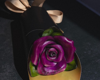 May include: A handcrafted, dark purple rose with green leaves emerges from a gold-colored, heart-shaped holder. The rose is part of a black, cylindrical package tied with a beige ribbon. The background is dark.