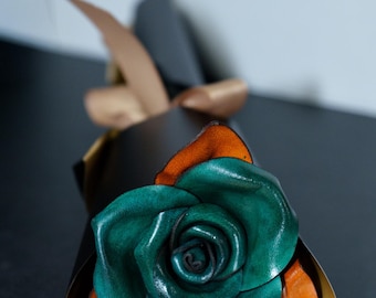 May include: A handcrafted leather rose featuring deep teal petals with visible orange undertones, wrapped in a dark brown paper cone secured with a light brown ribbon. The flower has a layered, organic structure with smooth, curved edges.