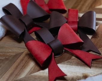 May include: Close-up of several handmade leather bows in shades of red and brown. The bows are arranged on a wooden surface with a chevron pattern. The bows have a classic bow shape, with a contrasting color on the front and back.