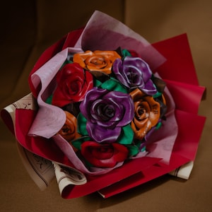 May include: A bouquet of handcrafted leather roses in vibrant shades of red, purple, and orange, accented with green leather leaves. The flowers are arranged together and wrapped in layers of deep red and light pink paper with printed text.