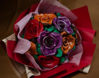 May include: A bouquet of handcrafted leather roses in vibrant shades of red, purple, and orange, accented with green leather leaves. The flowers are arranged together and wrapped in layers of deep red and light pink paper with printed text.