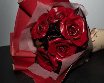 May include: A bouquet of deep red roses crafted from leather, featuring layered petals with dark brown edges. The flowers are arranged in a cluster and wrapped in layers of red and translucent white paper, creating a structured floral arrangement.