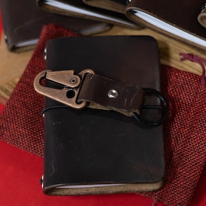 May include: A dark brown leather journal with a bronze-colored clasp and black ring closure. The journal is resting on a red surface, with other journals visible in the background. A small red pouch is also present.