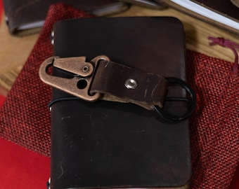 May include: A dark brown leather journal with a bronze-colored clasp and black ring closure. The journal is resting on a red surface, with other journals visible in the background. A small red pouch is also present.