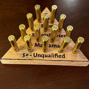 May include: A wooden triangle game board with brass casings. The board has the text "Expert", "Sharpshooter", "Marksman", and "5+ Unqualified" inscribed on it.