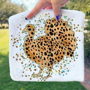 May include: White t-shirt featuring a turkey design with a leopard print pattern. The turkey is outlined in gold and surrounded by brown and teal paint splatters. The shirt is held up by a person.