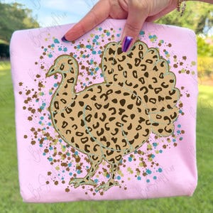 May include: A light pink t-shirt featuring a turkey design with a leopard print pattern. The turkey is outlined in gold and surrounded by brown and teal speckles. The shirt is held up by a person.