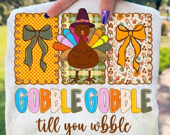 May include: White t-shirt with a Thanksgiving theme. The design includes three decorative squares with bows and a turkey wearing a pilgrim hat. The text "GOBBLE GOBBLE till you wobble" is printed in colorful letters.