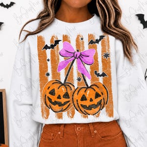 May include: White sweatshirt with a Halloween-themed design. The graphic features two orange jack-o'-lanterns with a purple bow, orange stripes, bats, and stars. The sweatshirt is paired with orange pants.