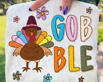 May include: White t-shirt featuring a colorful turkey wearing a pilgrim hat, with the word "GOBBLE" in vibrant letters. The design includes floral accents, perfect for Thanksgiving celebrations.