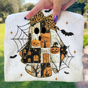 May include: White Halloween-themed square pillow featuring a patchwork ghost design with a leopard print bow. The ghost has pumpkin and house details, surrounded by bats and spiderwebs. The pillow is held up by a person.