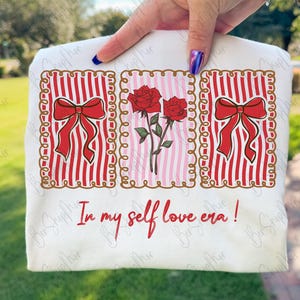 May include: A white t-shirt featuring three framed designs. The designs include red bows on red and white striped backgrounds and red roses on a pink and white striped background. The text "In my self love era!" is printed below.