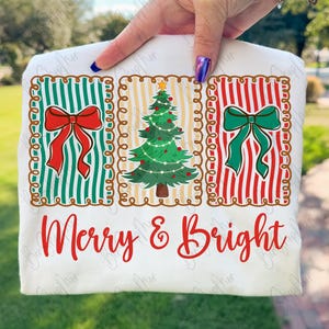 May include: White long-sleeve shirt with a Christmas-themed design. The design features three rectangular panels with a red bow, a Christmas tree, and a green bow. The words "Merry & Bright" are written in red.