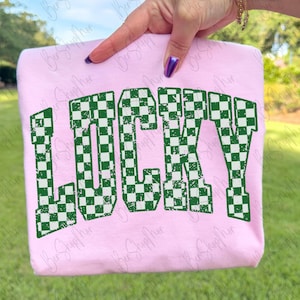 May include: A light pink sweatshirt with the word "LUCKY" in large, distressed, green and white checkered letters. The sweatshirt is held up by a hand with purple nail polish, against a background of green grass and trees.