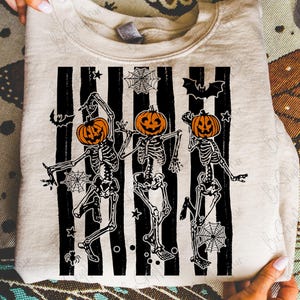 May include: A beige sweatshirt featuring a Halloween design. The graphic includes three skeletons with orange pumpkin heads, dancing in front of black and white stripes. Bats, spiderwebs, and stars complete the spooky theme.