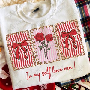 May include: White sweatshirt with a graphic design featuring red bows, roses, and the text "In my self love era!". The design is framed with a gold border. The sweatshirt is part of a holiday theme.