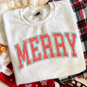 May include: A white crewneck sweatshirt featuring the word MERRY printed in a distressed red font with a green outline. The garment is displayed alongside red and green plaid fabric, a red stocking, and a plush reindeer toy.