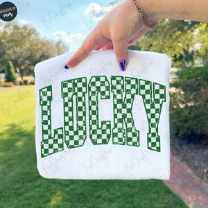May include: White sweatshirt with the word "LUCKY" in large, green, checkered letters. The sweatshirt is held up against a background of green foliage and a blue sky. The design is a bold, eye-catching statement.